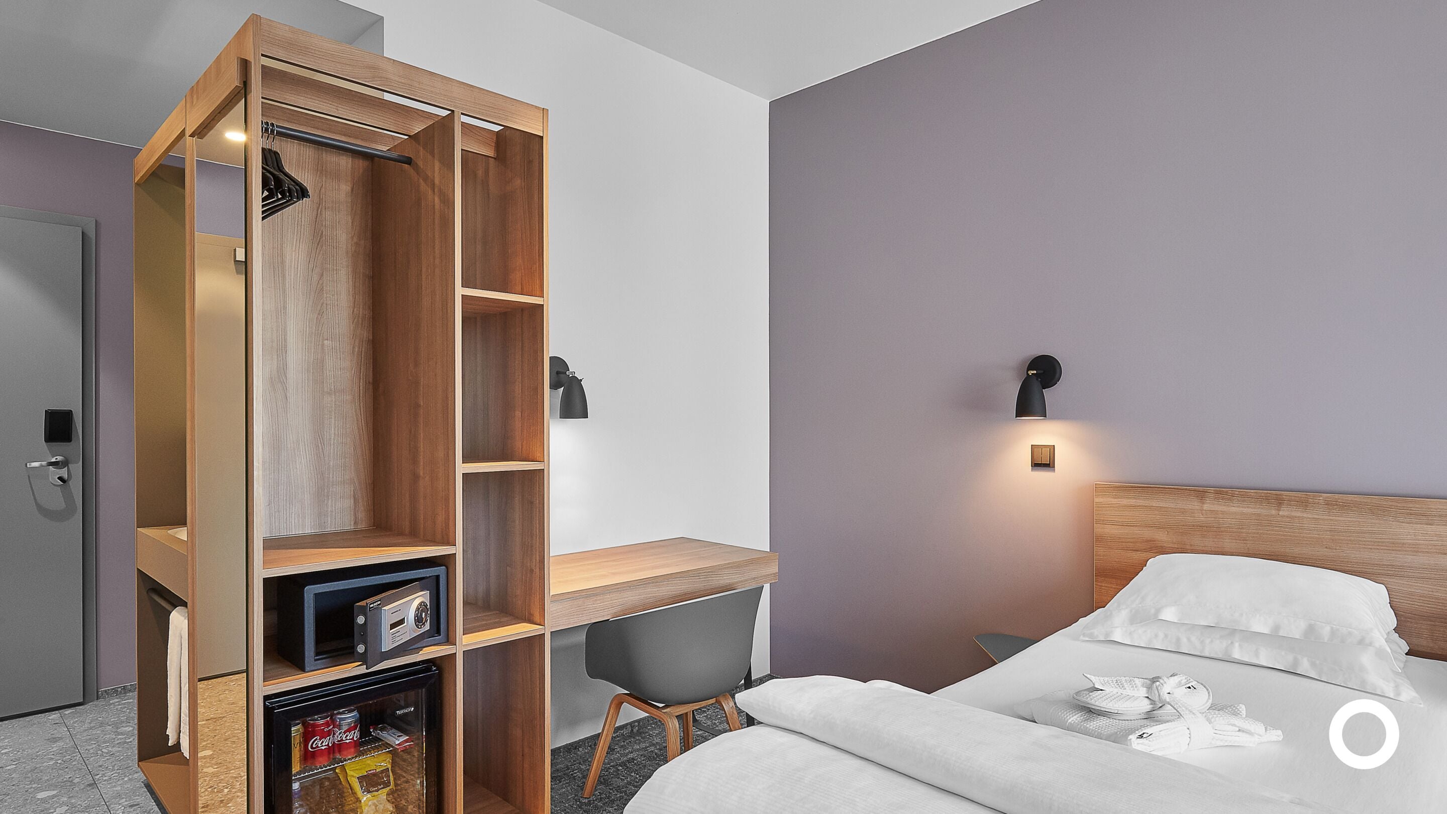 Una habitación de hotel con un escritorio de madera, una silla, una cama con ropa de cama blanca y una lámpara fijada a la pared.