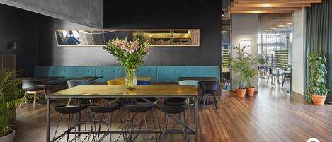 Un comedor moderno con una mesa larga, múltiples sillas y un jarrón de flores. Hay plantas en macetas alrededor de la habitación, y una zona de estar con un banco tapizado en azul.