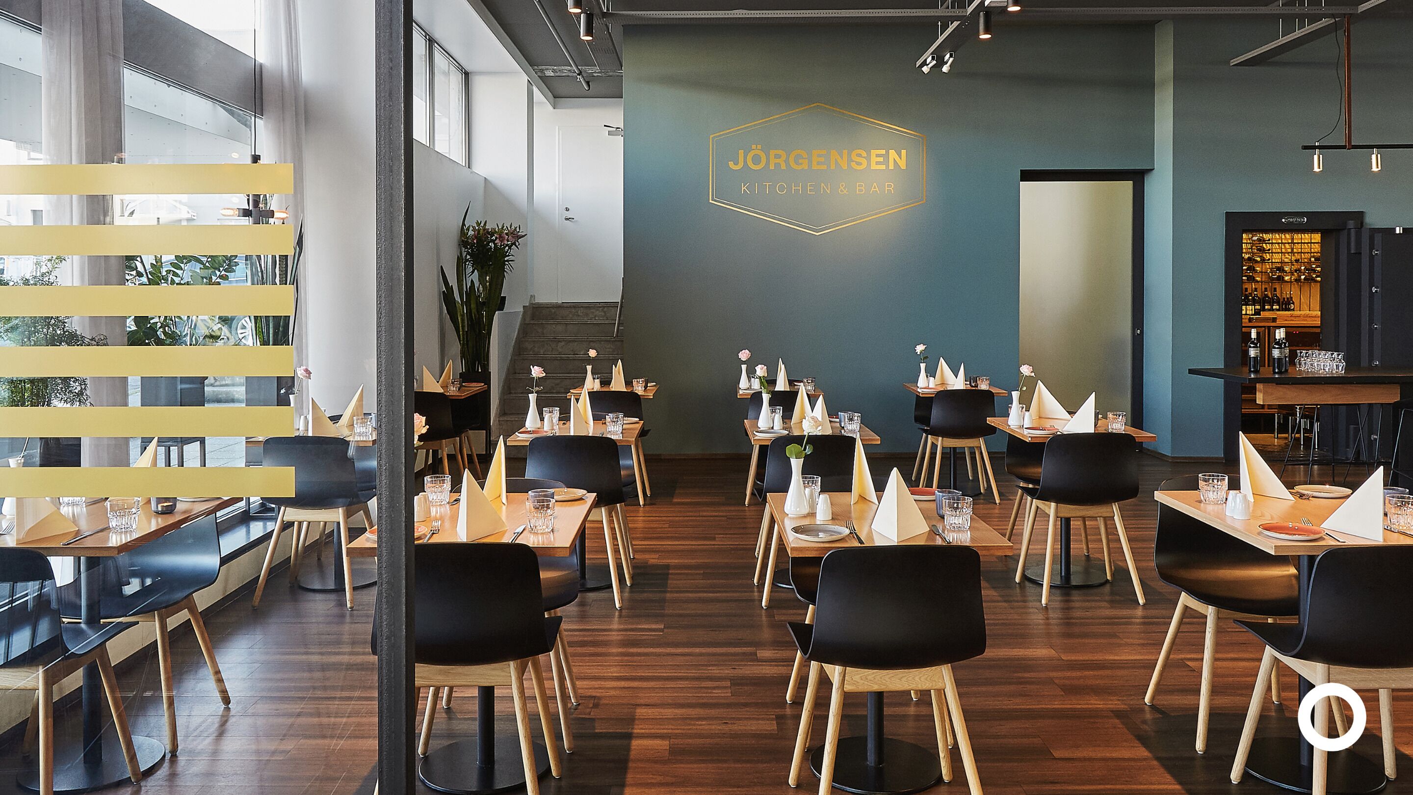A modern restaurant interior with a central bar area, wooden tables, black chairs, and a neon sign reading "JÖRGENSEN KITCHEN & BAR".