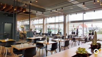Un café moderno con mesas y sillas de madera, ventanas amplias y plantas de interior.