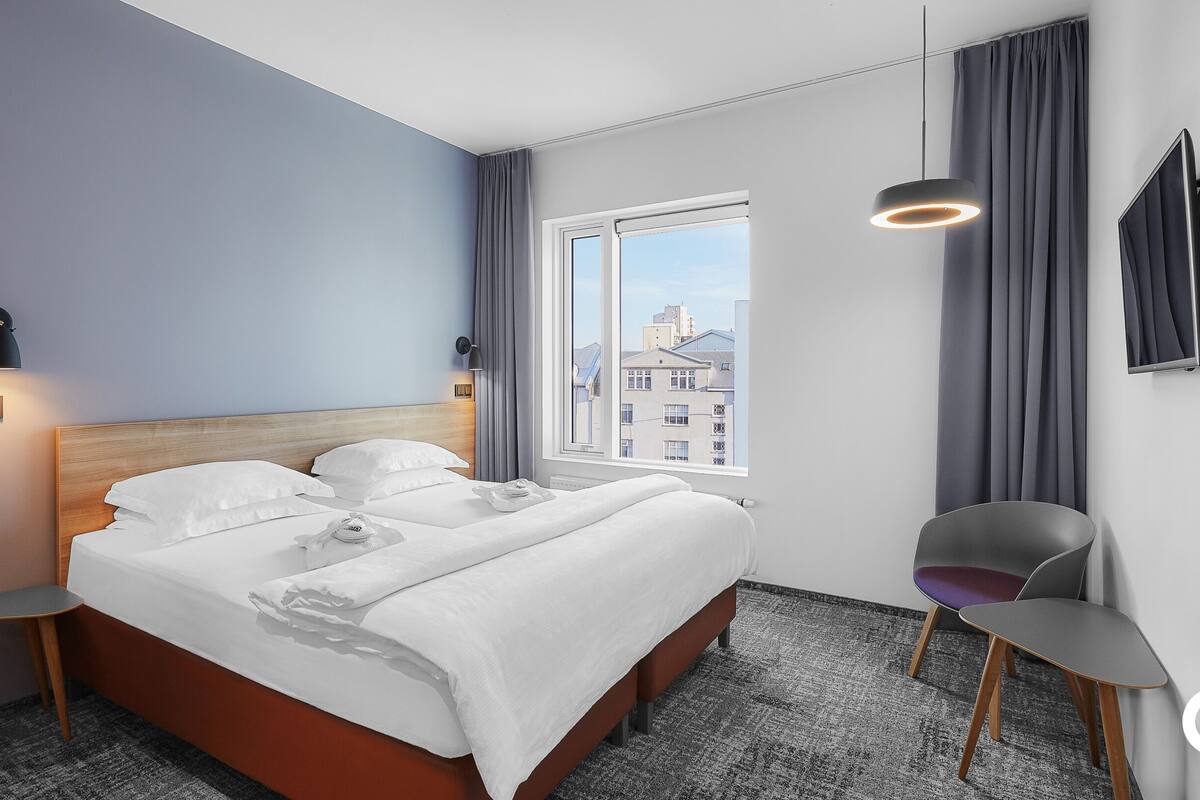 Et moderne hotelværelse med en stor seng, et fjernsyn, et skrivebord og udsigt til bygninger gennem vinduet.