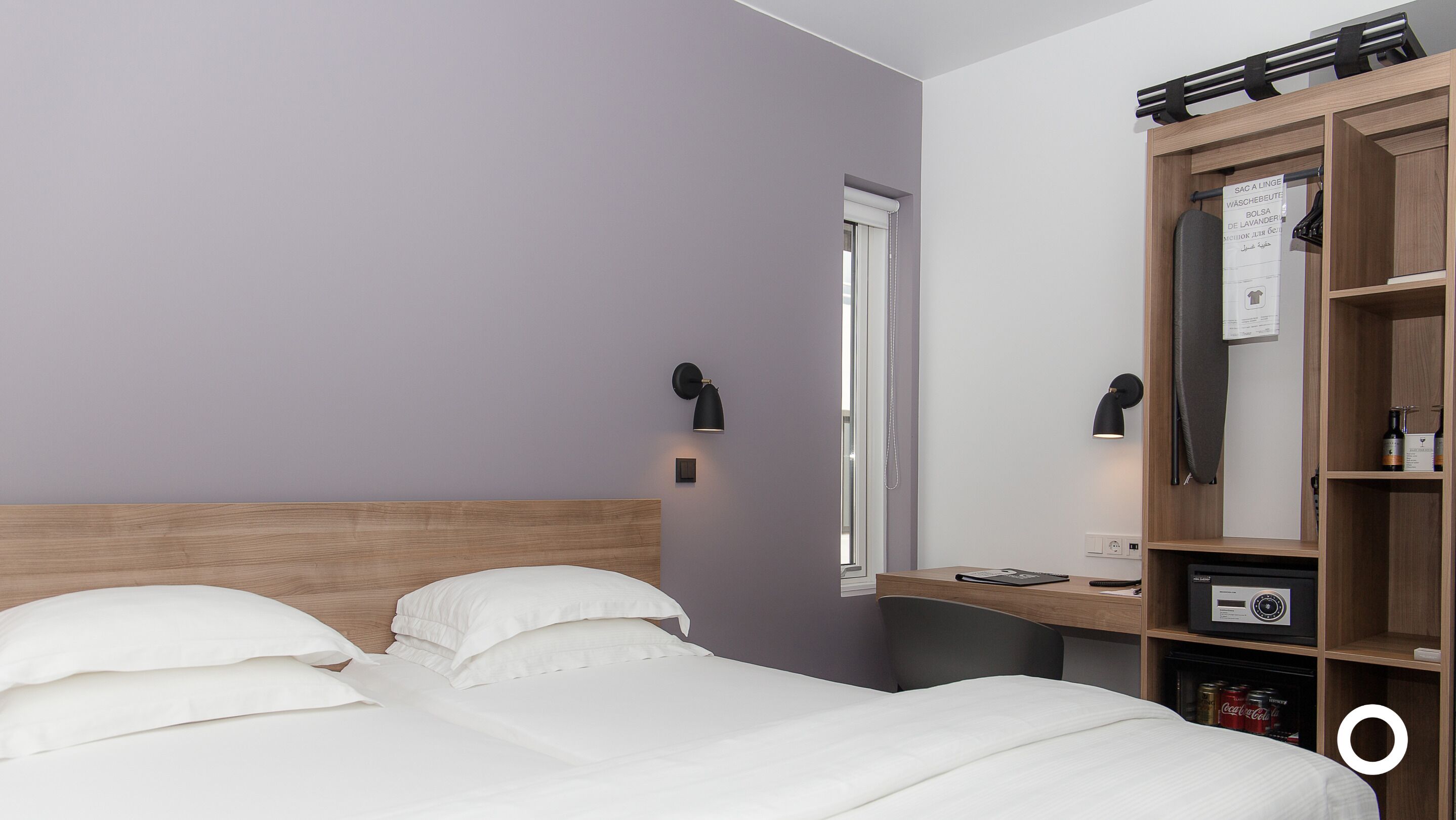 A hotel room with a bed, two wall-mounted lamps, a wooden desk, a mirror, and a wardrobe with a hanging rack.