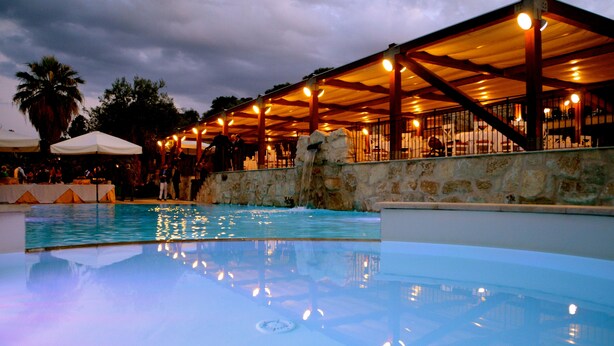 A swimming pool with a waterfall feature, surrounded by a covered seating area with tables and chairs. - Tenuta di Roccadia (Carlentini)