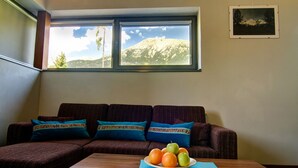 Een woonkamer met een bank, een raam met uitzicht op de bergen, en een tafel met een fruitschaal.