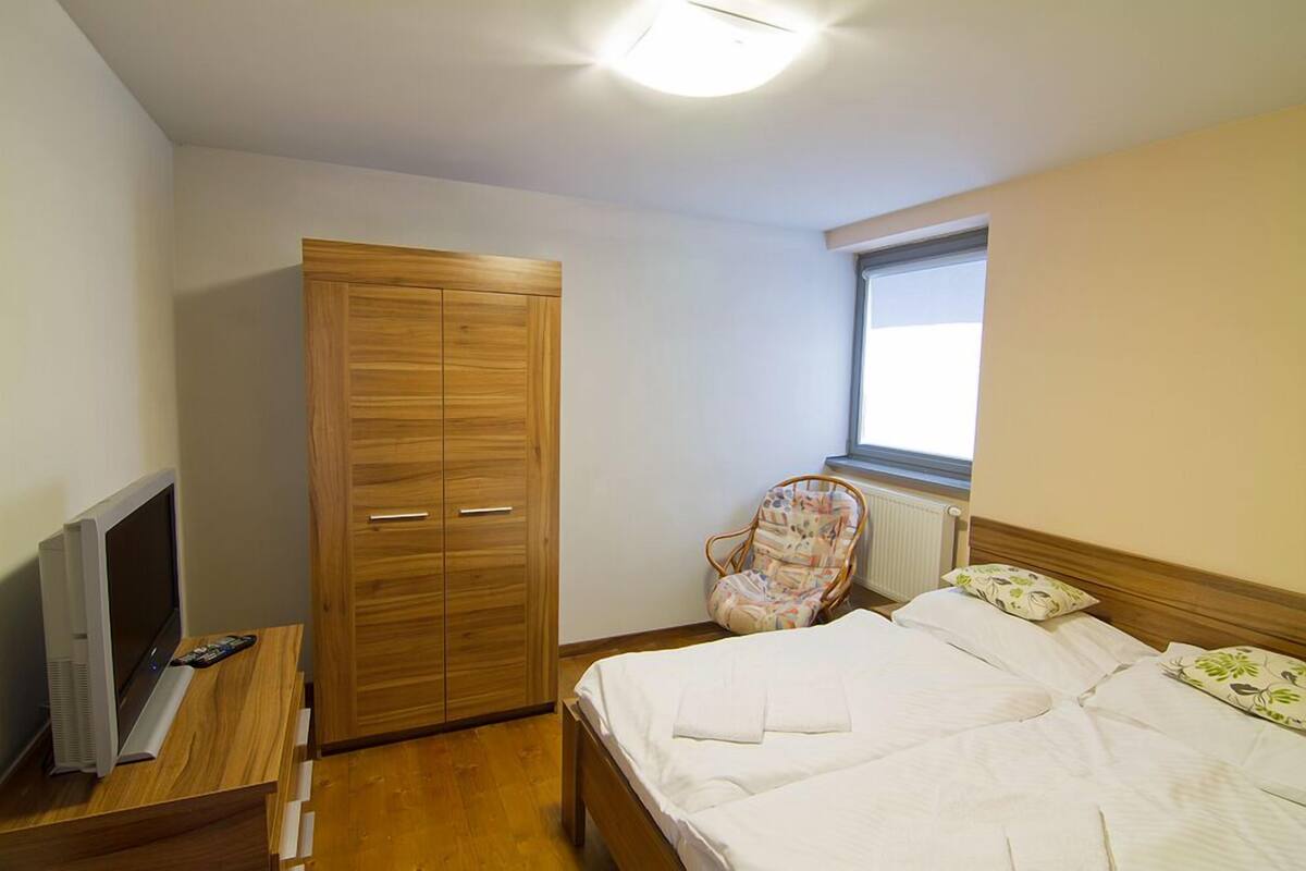 Ein Schlafzimmer mit einem Holzschrank, einem Bett mit weißer Bettwäsche, einem Sessel, einem Fernseher auf einem Holztisch und einem Fenster mit Jalousien.