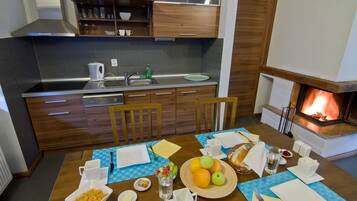 A modern kitchen with wooden cabinets, a stainless steel sink, and a built-in oven. There is a dining table set with plates, cups, and a fruit bowl.