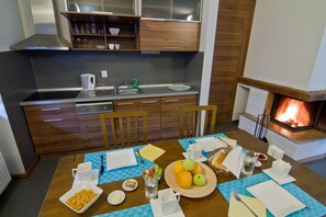 A modern kitchen with wooden cabinets, a stainless steel sink, and a built-in oven. There is a dining table set with plates, cups, and a fruit bowl.
