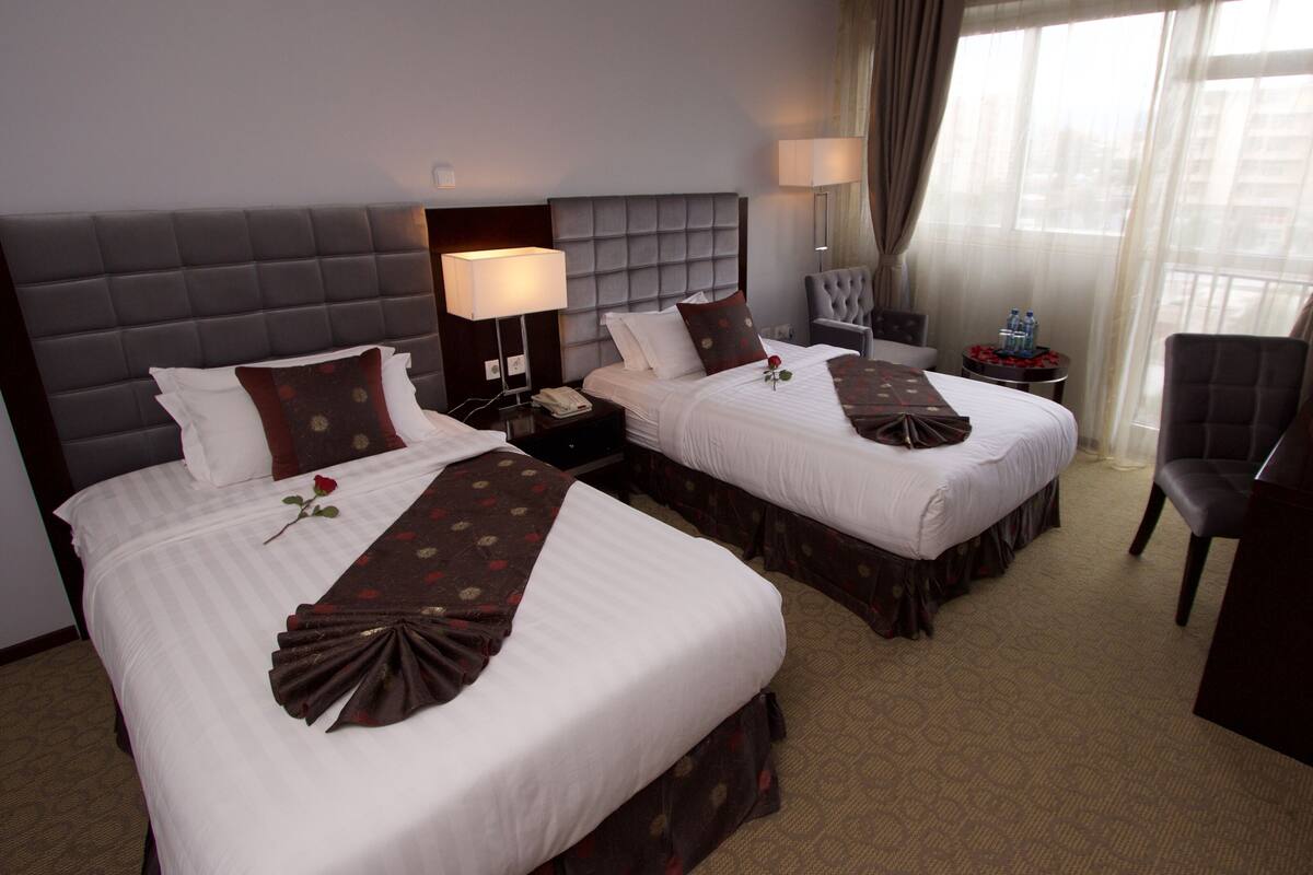 A hotel room with two beds, a desk, a chair, and a view of the city through the window.