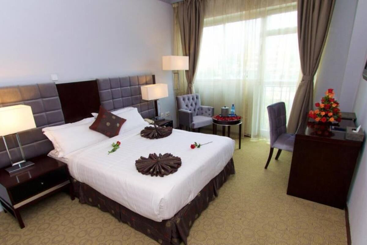 A hotel room with a large bed, bedside lamps, a desk with a chair, a TV, and a small table with flowers.
