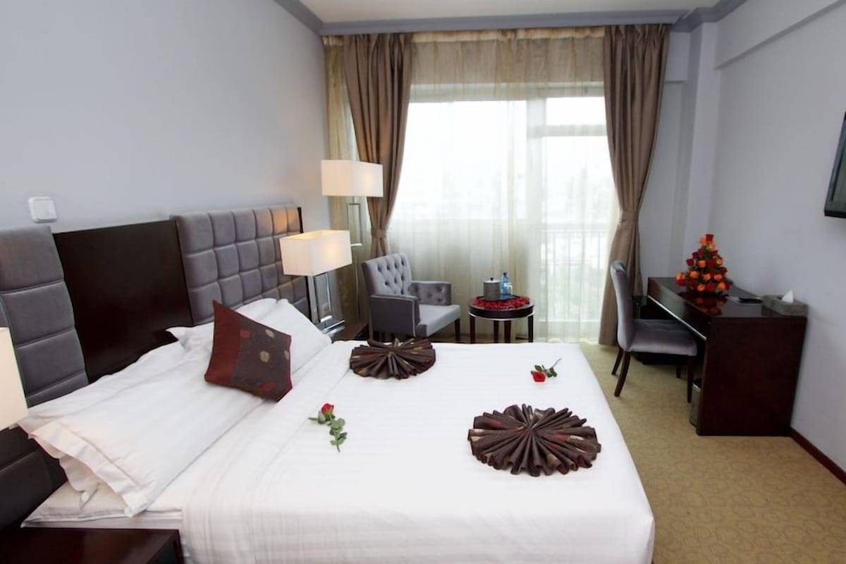 Habitación de hotel con cama, escritorio, silla, televisor y ventana con cortinas.