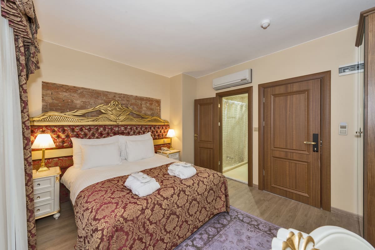 A hotel room with a large bed, a headboard with a decorative design, a bedside table with a lamp, and a wardrobe.