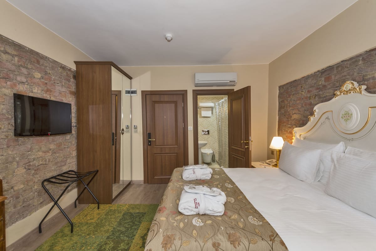 A hotel room with a brick wall, a flat-screen TV, a bed with white linens, and a wooden wardrobe.