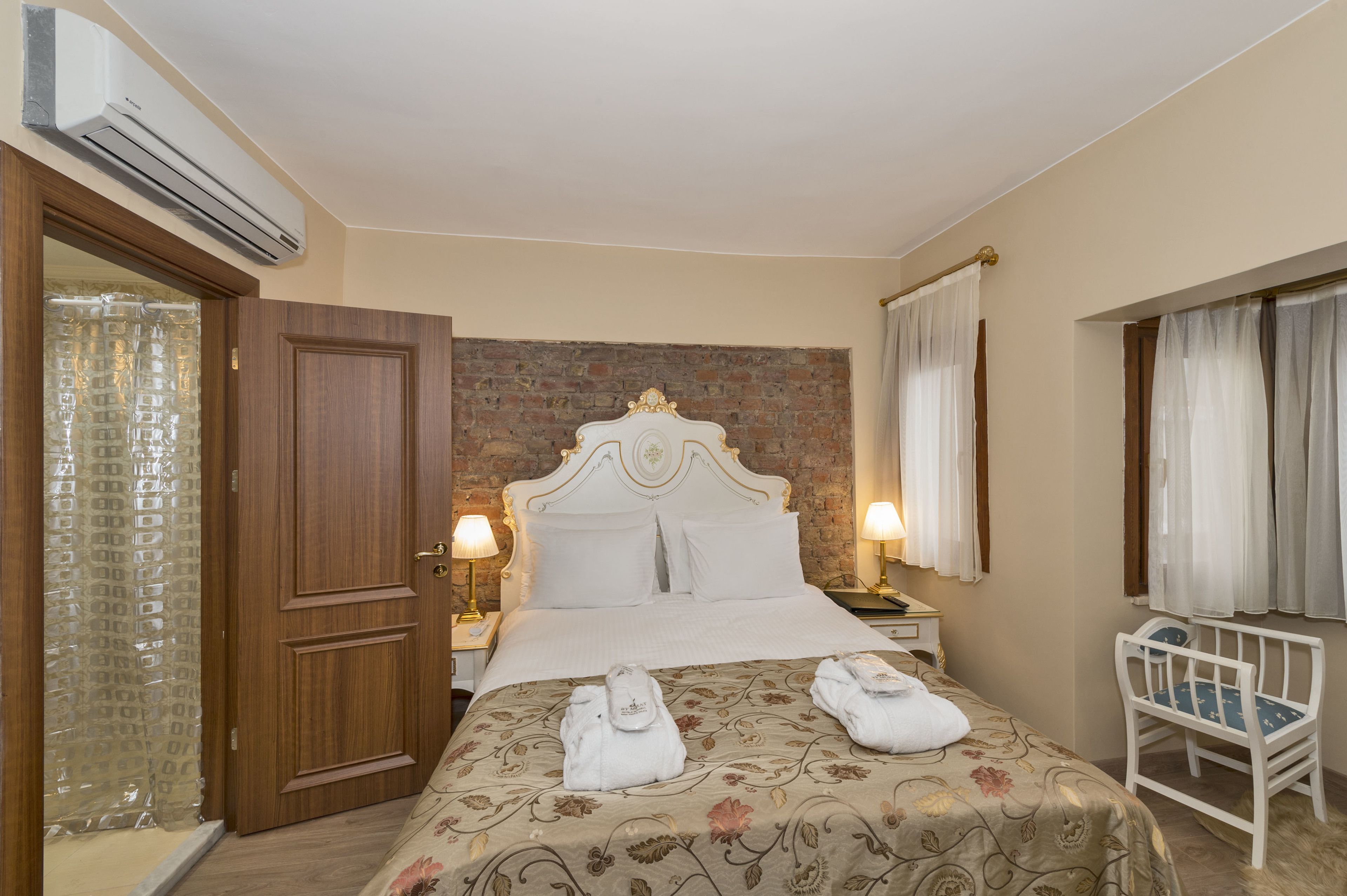 A hotel room with a wooden door, a bed with a floral bedspread, two bedside lamps, a white headboard, and a window with curtains.