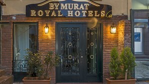Bymurat Royal Hotels har en mörk entrédörr och en fasad som pryds av väggmonterade lampor och krukväxter vid ingången.