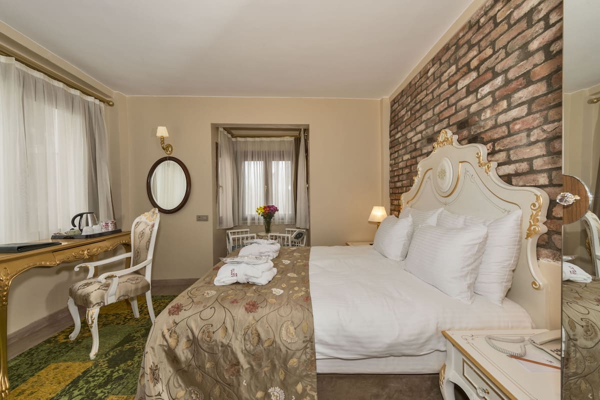 A hotel room with a bed, a desk with a chair, a mirror, and a brick wall.
