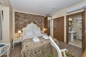 A hotel room with a brick accent wall, a large bed with a floral bedspread, two bedside lamps, a white chair, and a bathroom with a sink and mirror. - By Murat Royal Hotels (Istanbul)