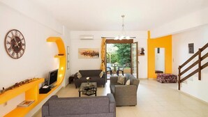 Ein Wohnzimmer mit einer Couch, einem Couchtisch, einem Fernseher und einem Kamin. Eine Treppe befindet sich rechts und eine Tür führt auf einen Balkon.