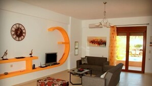 Ein Wohnzimmer mit Sofa, Couchtisch und Fernseher auf einem Schrank. An der Wand befindet sich ein großes, oranges 'C', eine Uhr und ein Fenster mit Vorhängen.