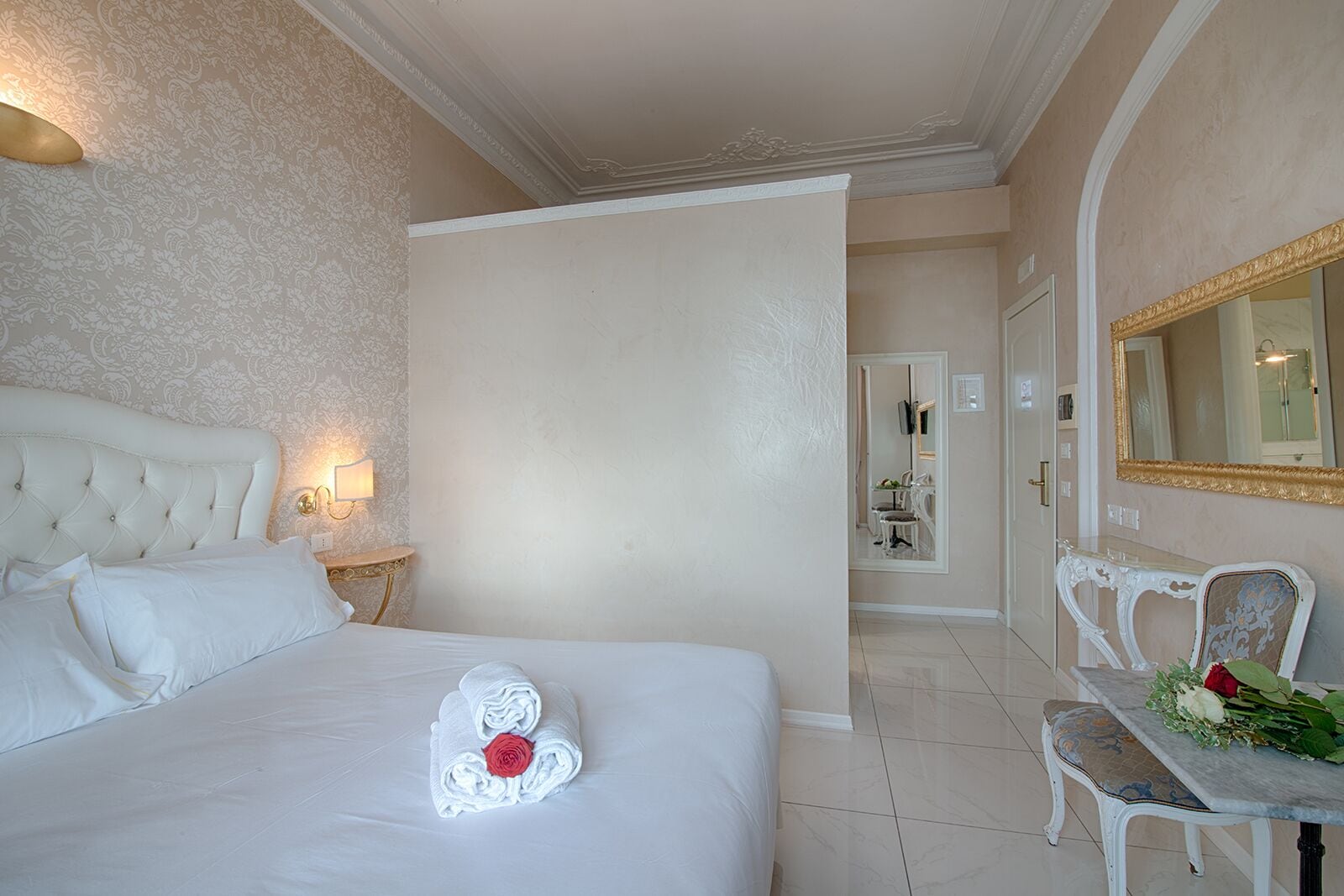 Una camera d'albergo con un letto grande, un'area salotto con tavolo e sedie, uno specchio e un bagno visibile attraverso una porta aperta.