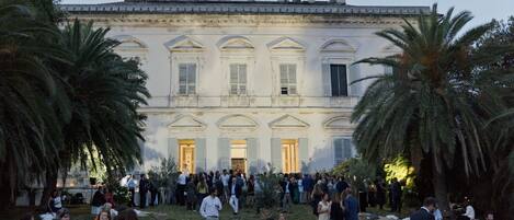 Evento al aire libre con un edificio grande al fondo, palmeras y personas reunidas alrededor de mesas.