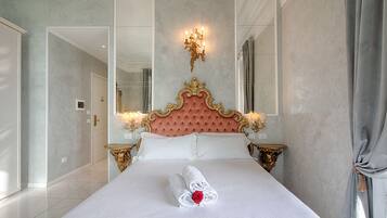Ein großes Bett mit geblattetem Kopfteil, zwei Nachttische mit goldfarbenen Akzenten, ein Spiegel über dem Bett und ein Fenster mit Vorhängen.