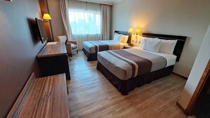 Quarto de hotel com duas camas, uma escrivaninha, uma cadeira e uma janela grande com cortinas.