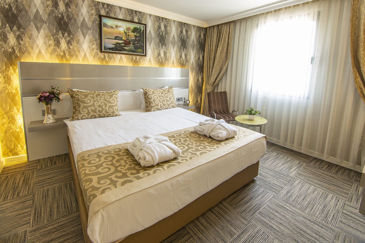 Habitación de hotel con una cama grande, cabecera, mesita de noche con una planta y una ventana con cortinas.