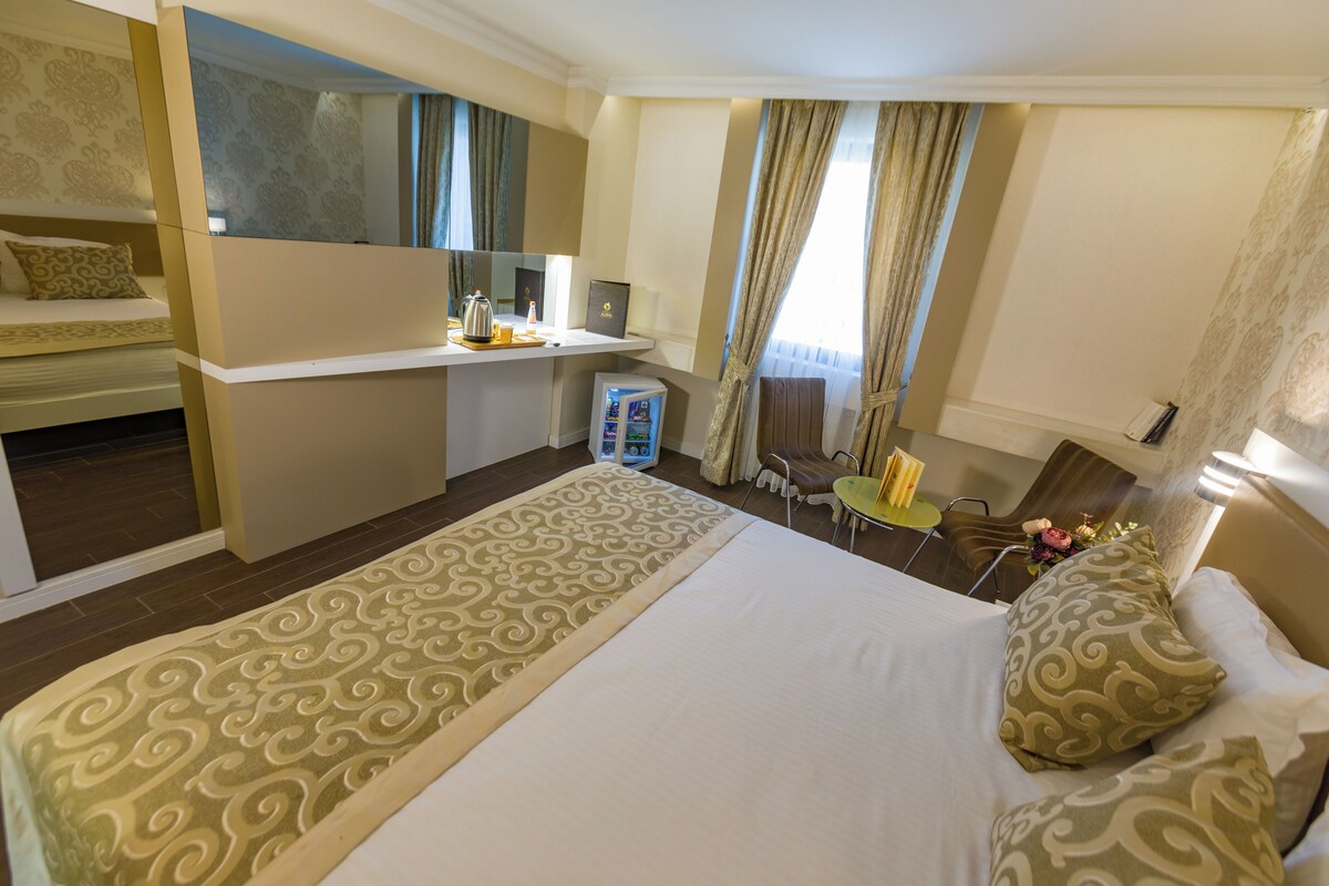 A hotel room with a large bed, a desk with a computer, a chair, and a small table with a vase of flowers.