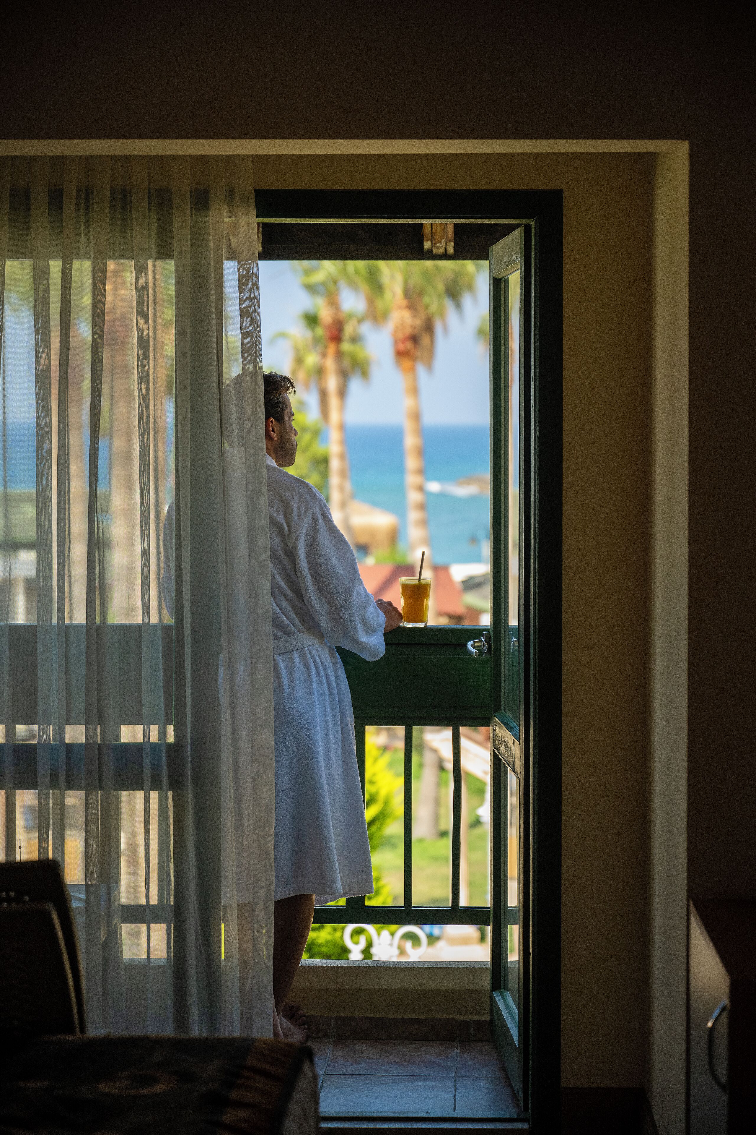 A person in a white bathrobe standing at a balcony door, looking out at a view of the ocean and palm trees.
