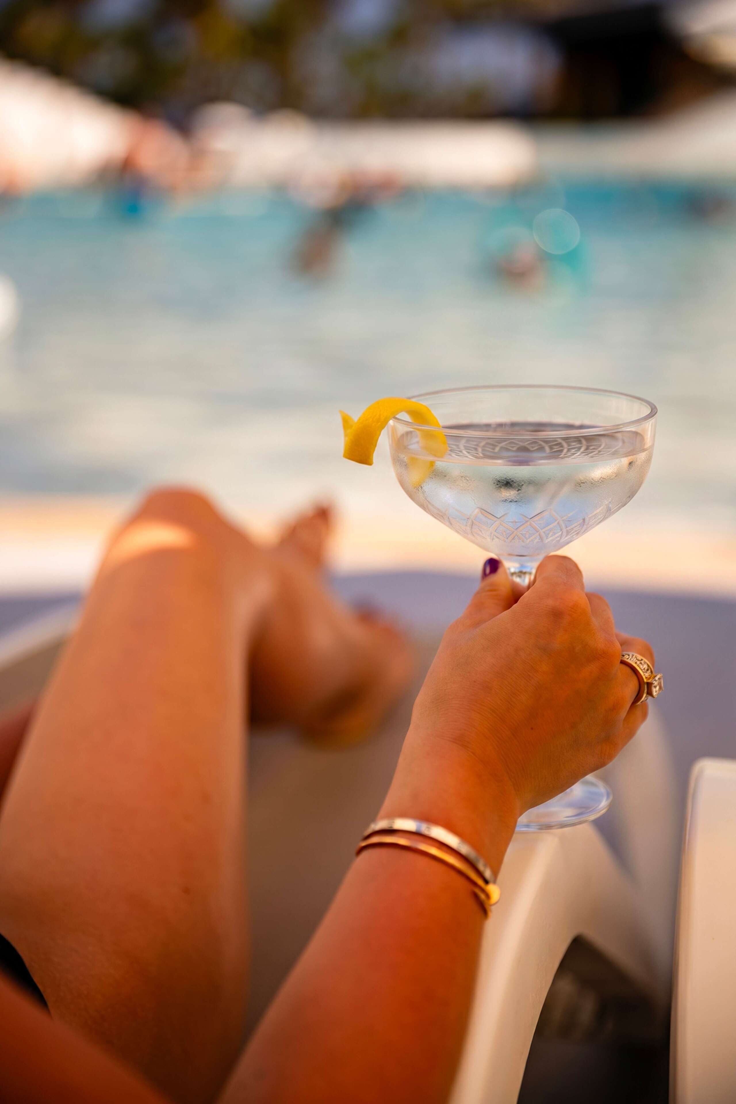 A person holding a clear glass with a lemon garnish, seated by a pool with a blurred background.