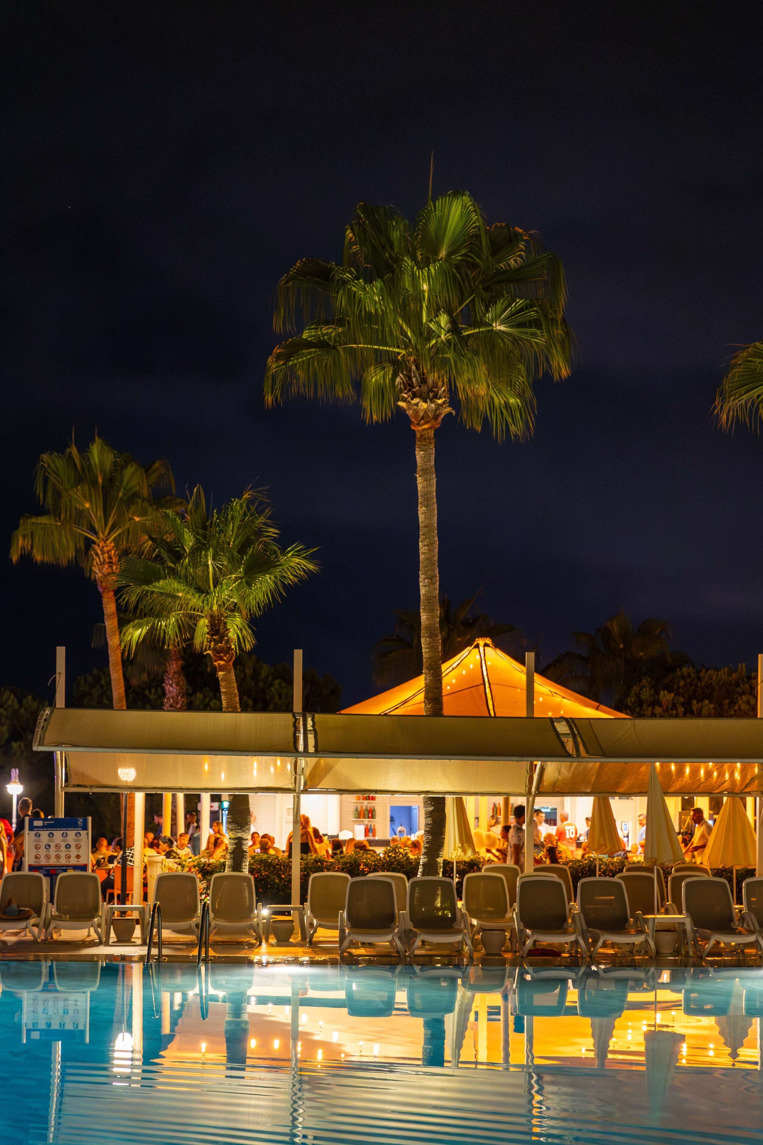 A pool area with lounge chairs, palm trees, and a building with a tent-like structure.
