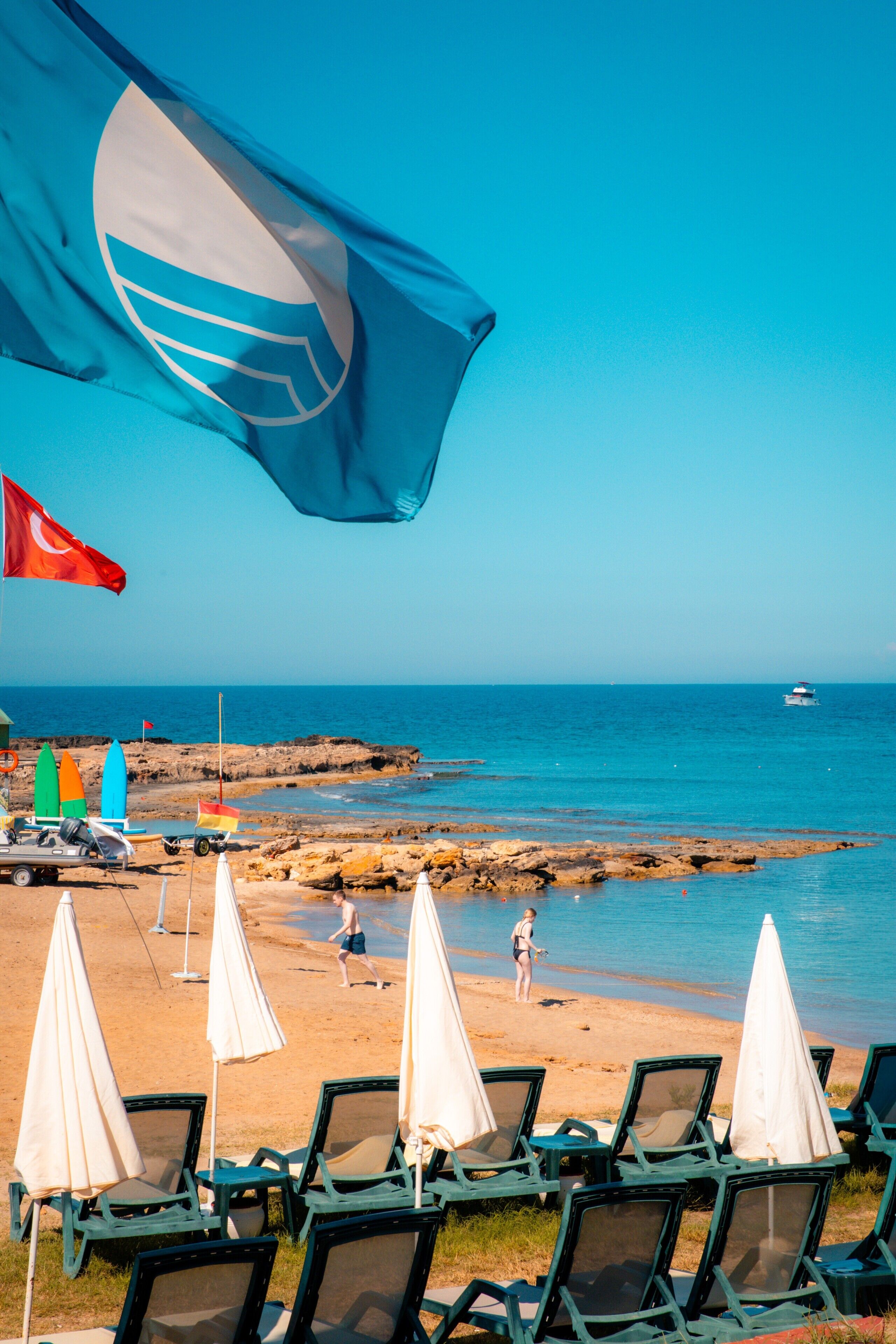A beachside view with lounge chairs and umbrellas, a flag, and a rocky shoreline.