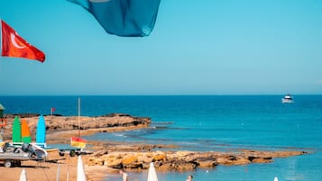 A beachside view with lounge chairs and umbrellas, a flag, and a rocky shoreline.