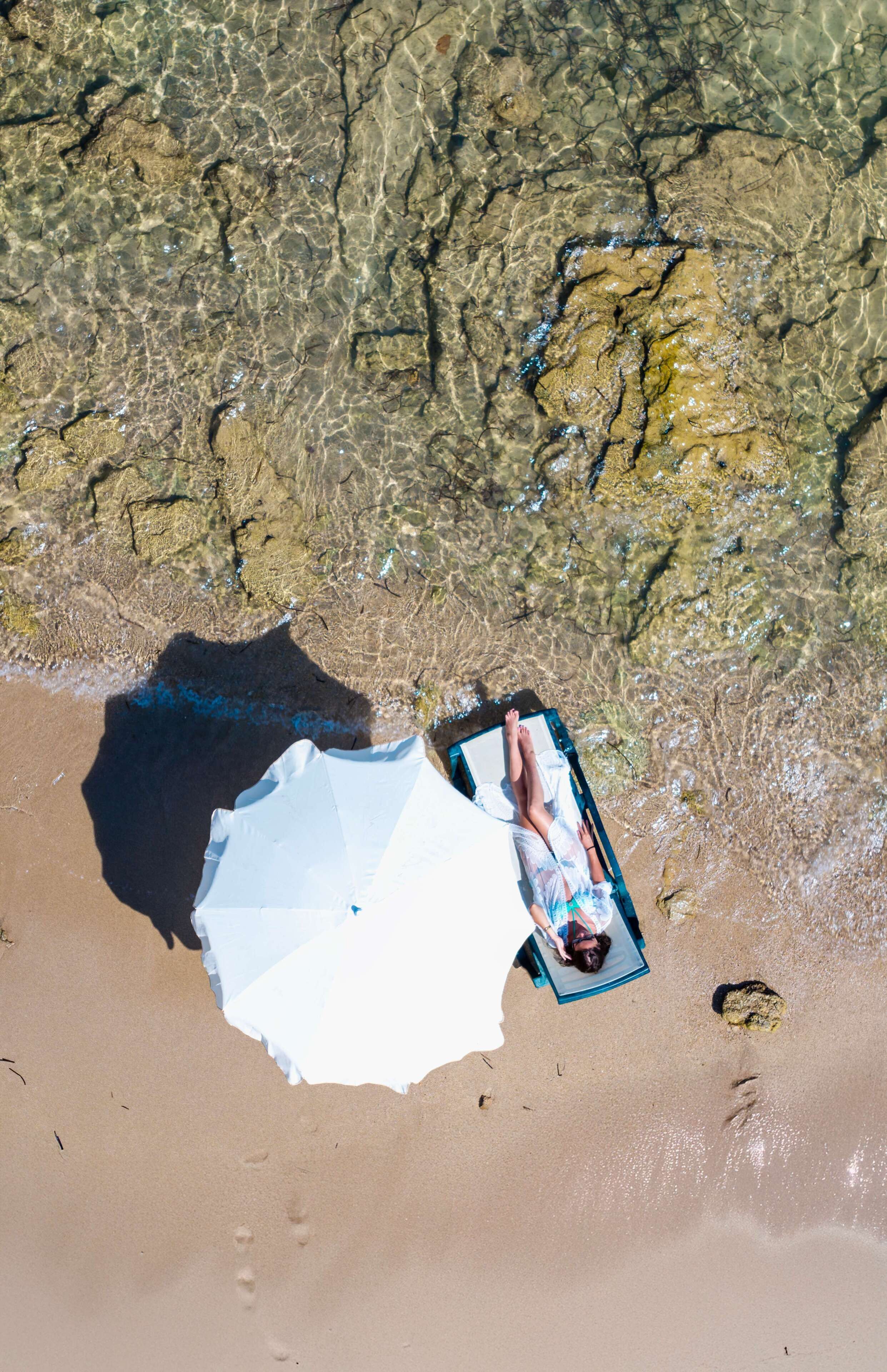 En strandscen med klart vatten, en person som ligger på en solstol och ett vitt parasoll.