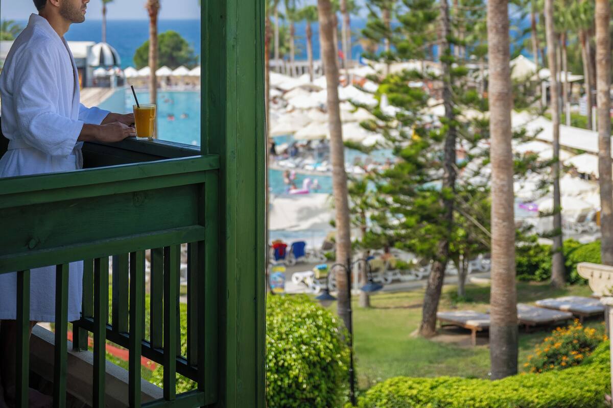 Ein Mann in einem weißen Hemd steht auf einer grünen Veranda mit Blick auf einen Poolbereich, Palmen und den Ozean.