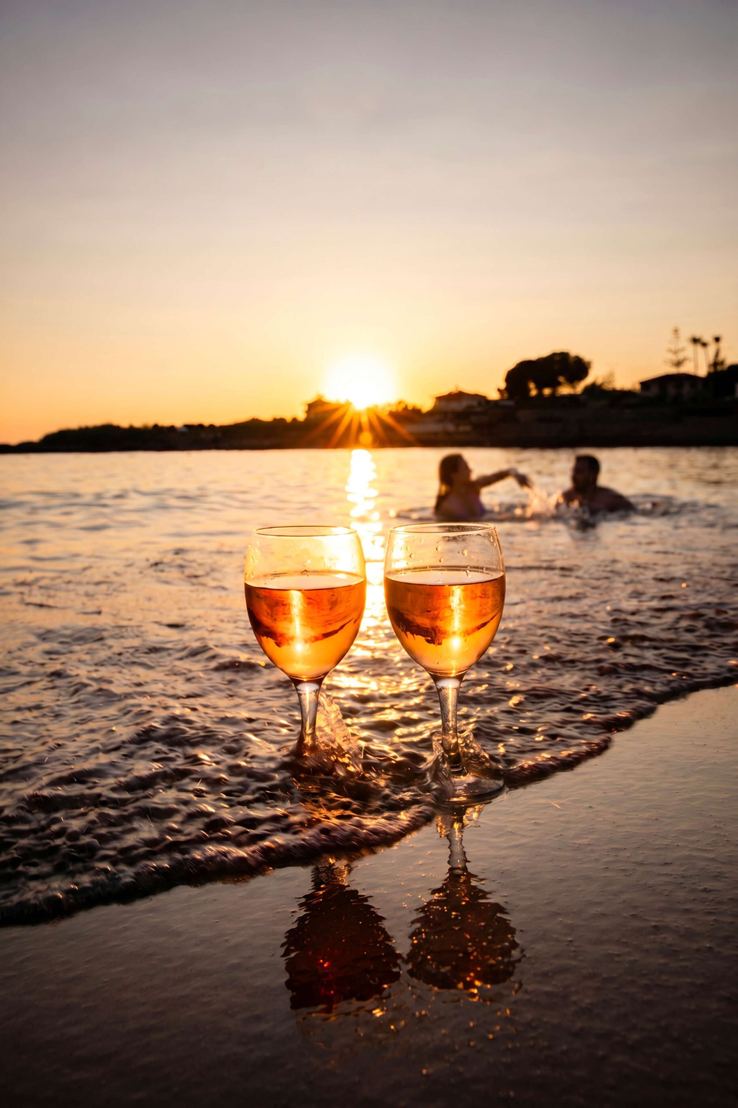 Two glasses of a clear liquid on a reflective surface, with a sunset and silhouettes of people in the background.
