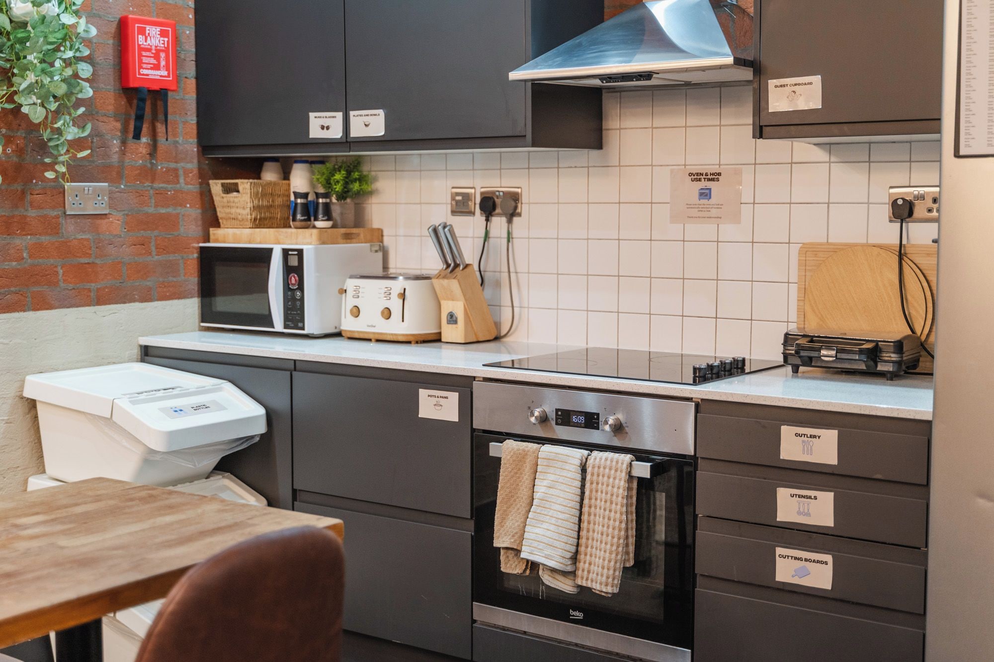 A modern kitchen with a microwave, toaster, and oven. There are cabinets labeled for pots & pans, cutlery, and utensils. A fire blanket is mounted on the wall.