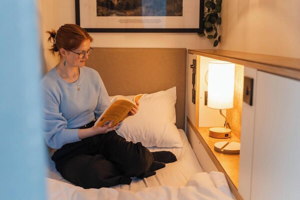 A person sitting on a bed reading a book in a room with a bedside lamp and a framed picture on the wall.