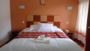 Una cama bien hecha con sábanas blancas, un plaid decorativo y una toalla doblada. Hay una mesita de noche con un teléfono y un control remoto. La habitación tiene una pared naranja y un cabecero de madera.