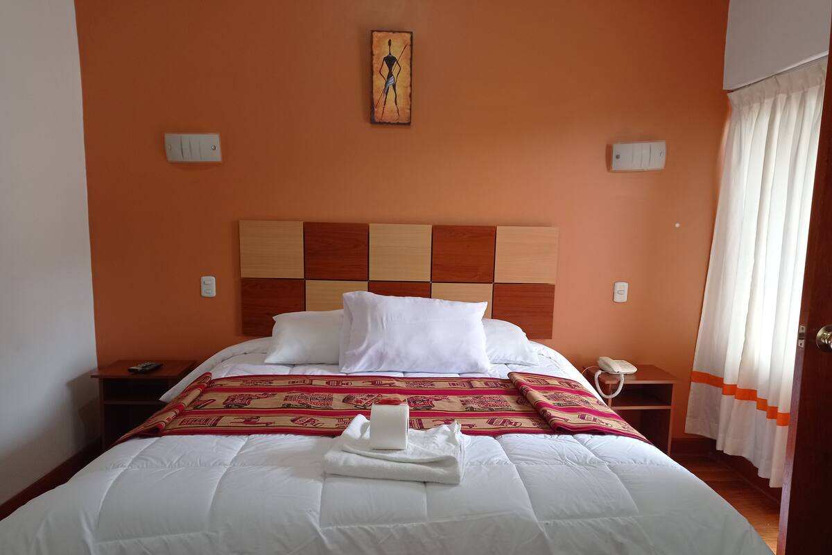 A neatly made bed with white linens, a decorative throw blanket, and a folded towel. There is a bedside table with a phone and a remote control. The room has an orange wall and a wooden headboard.