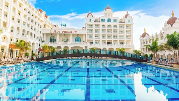 A large swimming pool with a bridge, surrounded by a hotel with multiple balconies and a sign reading "SIDE PREMIUM". - Oz Hotels Side Premium (Manavgat)