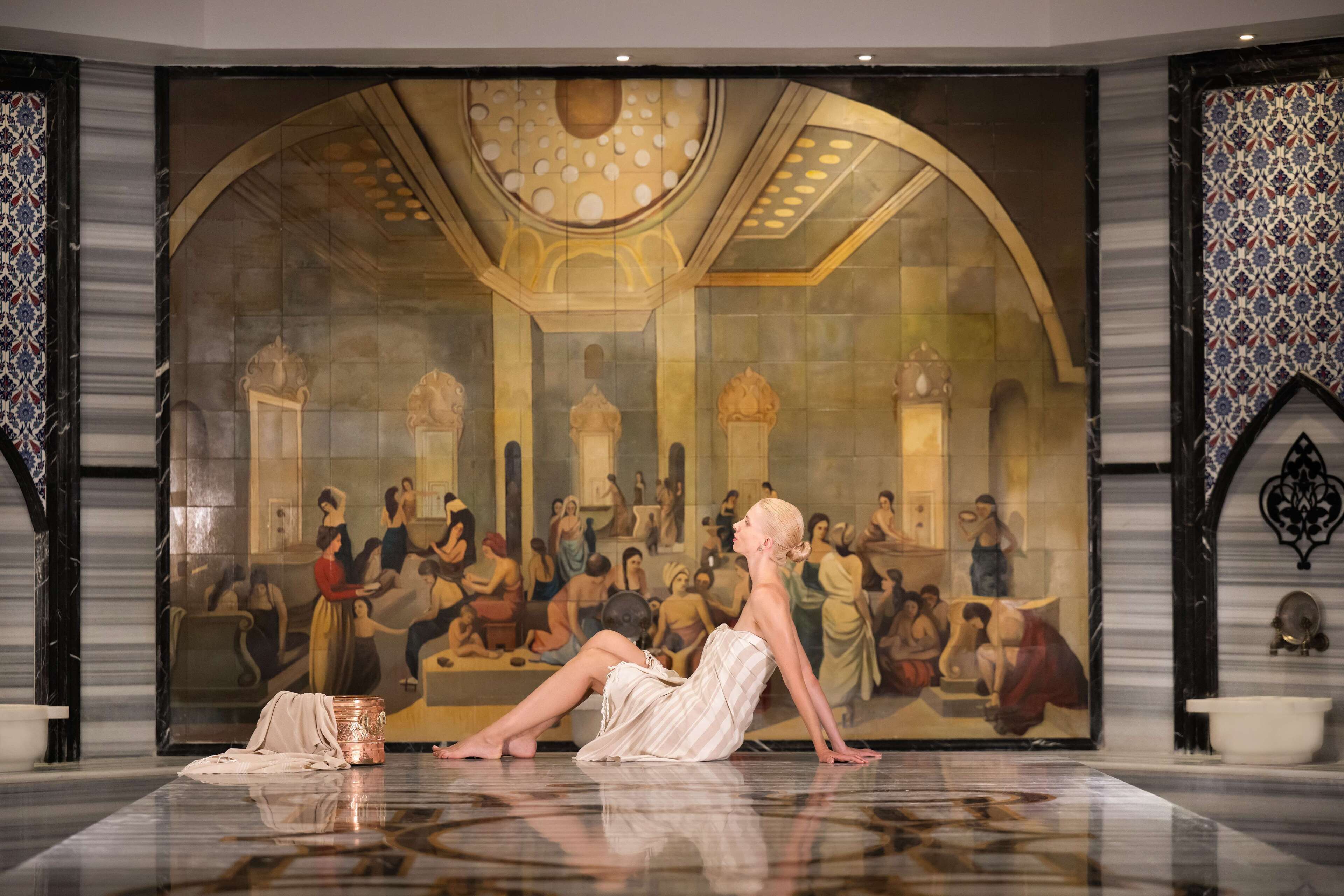 A person performing in front of a large mural in a room with a reflective floor and ornate decor.