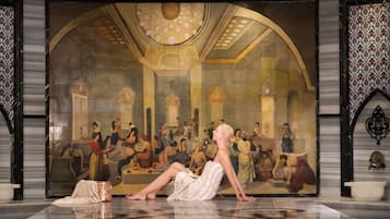 A person performing in front of a large mural in a room with a reflective floor and ornate decor.