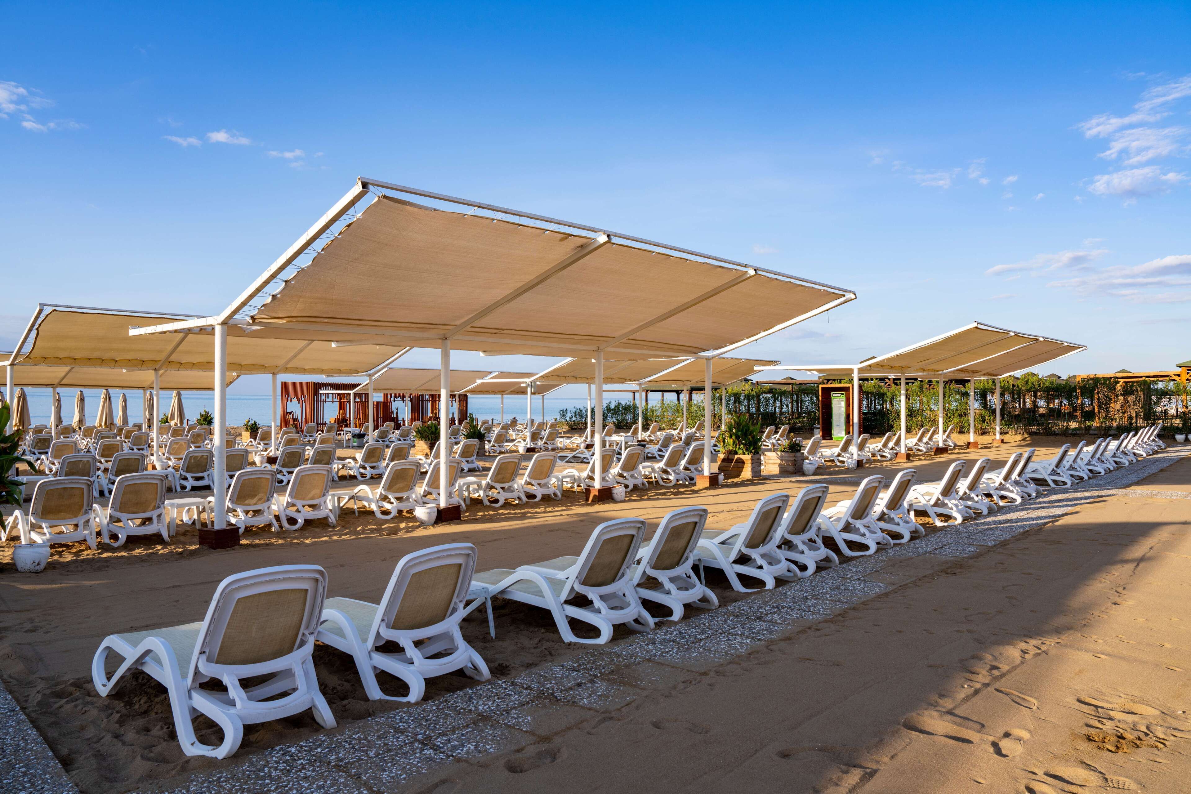 Eine Strandlounge mit Reihen weißer Liegen unter großen Sonnensegeln, einem Sandweg und freiem Blick auf das Meer.