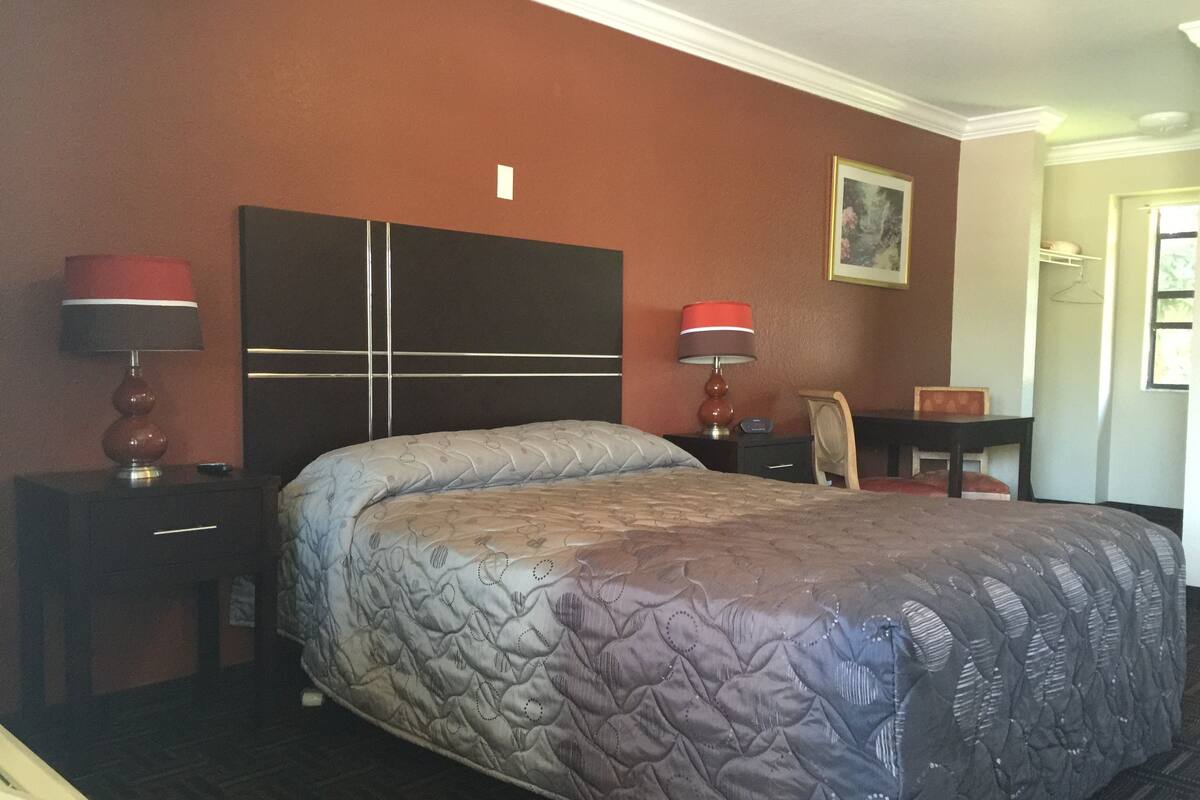 Ein Hotelzimmer mit einem großen Bett, zwei Nachttischen mit Lampen, einem kleinen Schreibtisch, einem Sessel und einem Bild an der Wand.