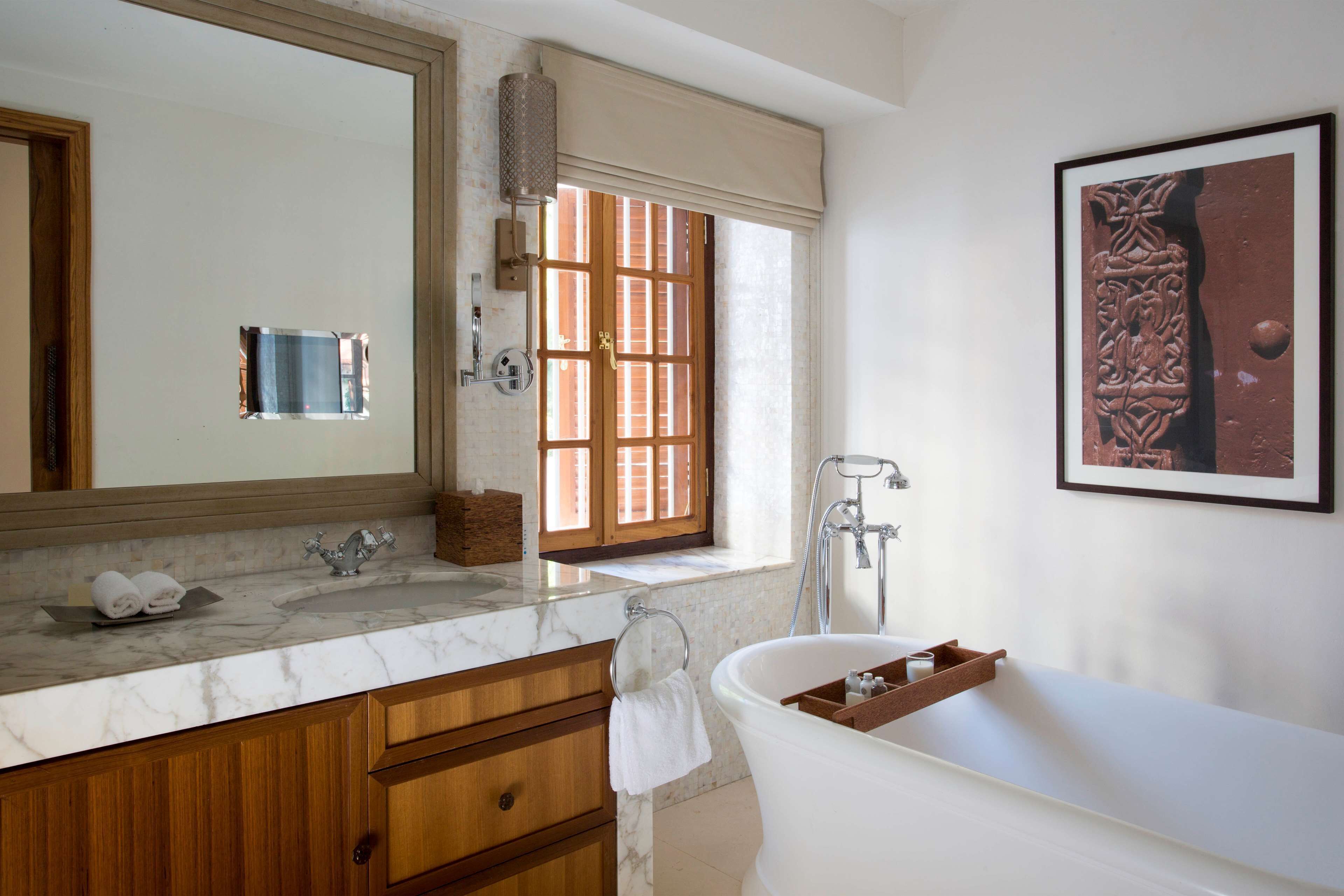 Ein Badezimmer mit Marmorplatte, Holzschränken, einem großen Spiegel, einem Fenster mit Läden, einer Badewanne und einem gerahmten Kunstwerk an der Wand.
