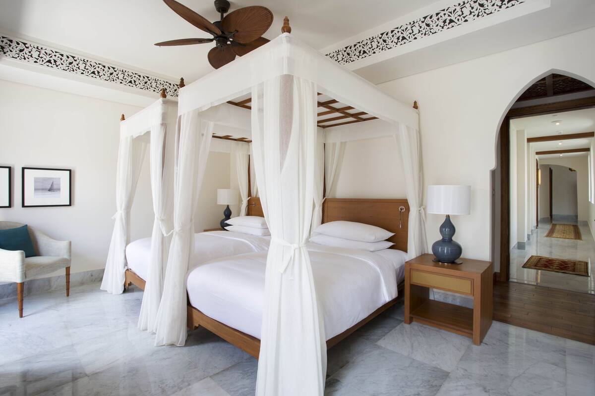 A four-poster canopy bed with white linens, a wooden headboard, and a nightstand with a lamp.