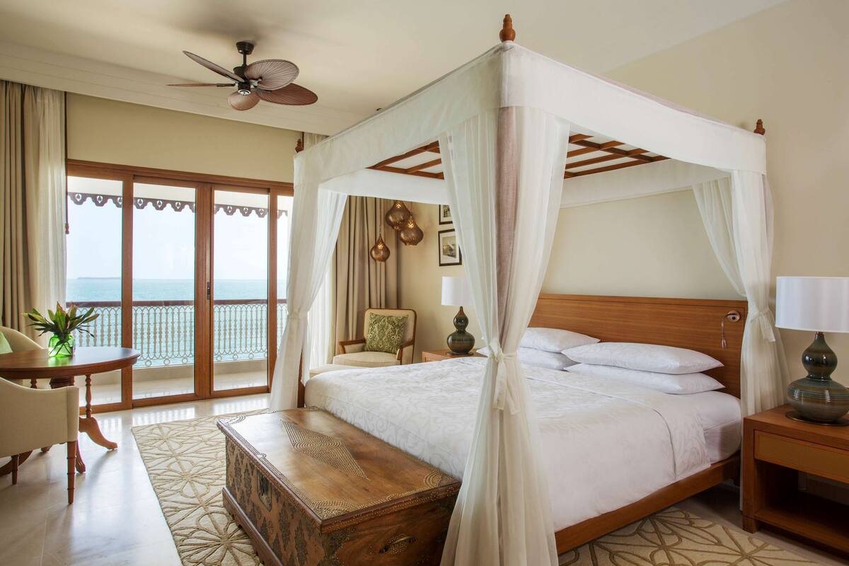 A four-poster canopy bed with a wooden headboard, a bedside table with a lamp, a wooden chest, and a view of the sea through a balcony with curtains.