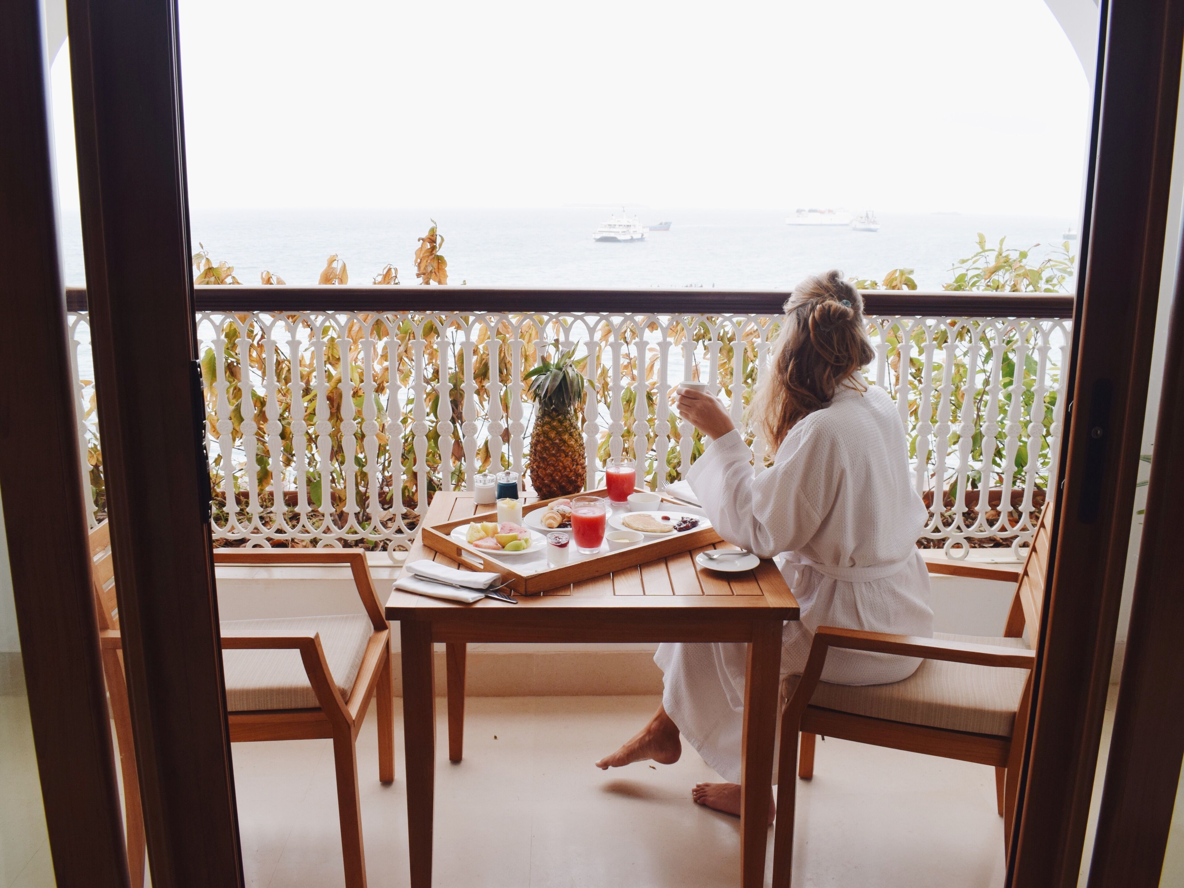 Une personne est assise à une table en bois, devant un plateau rempli de nourriture et de boissons. Elle est face à un balcon d’où l’on peut admirer la mer et les bateaux.