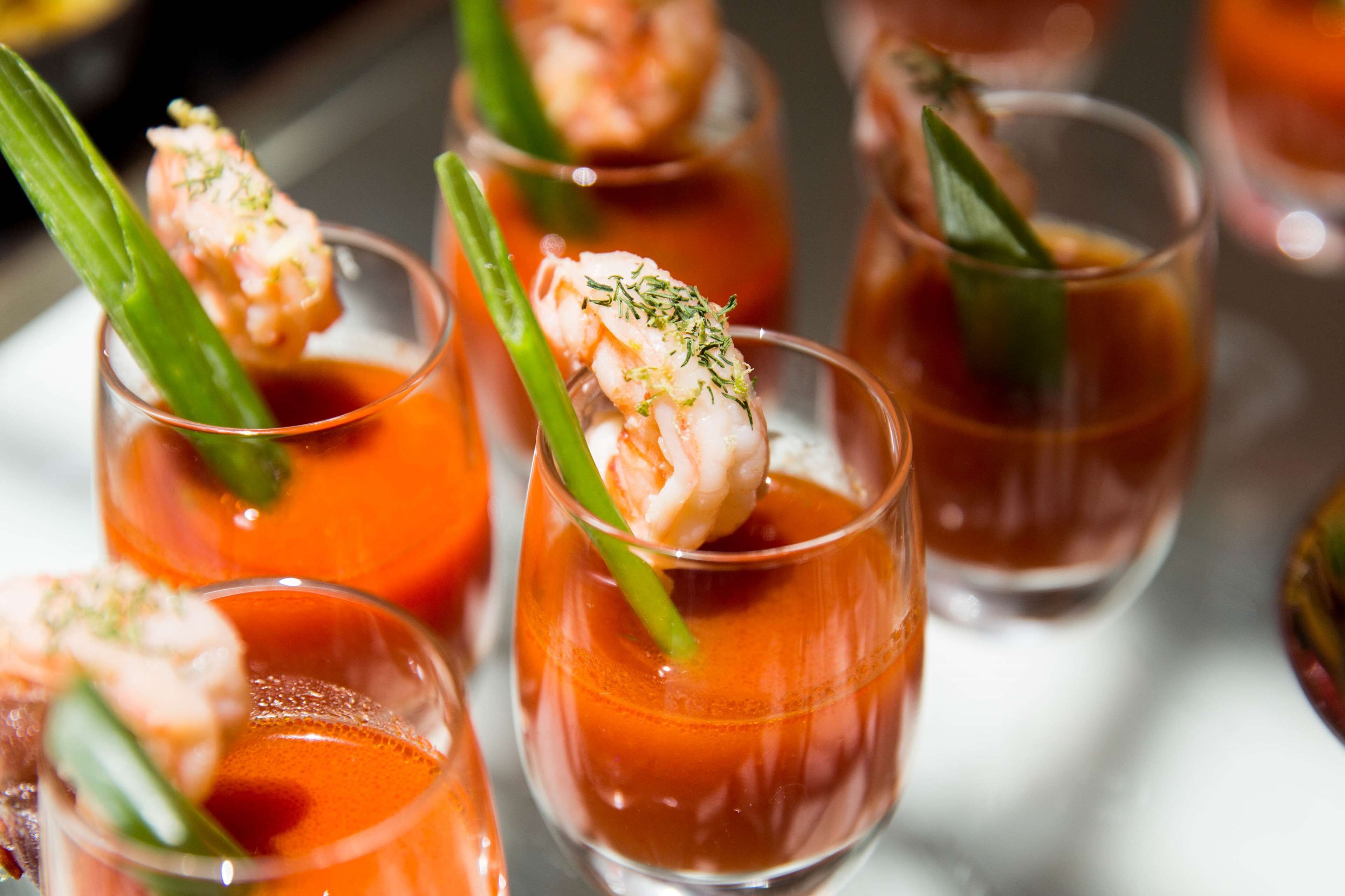A series of clear glasses containing a red liquid, garnished with shrimp and green herbs.